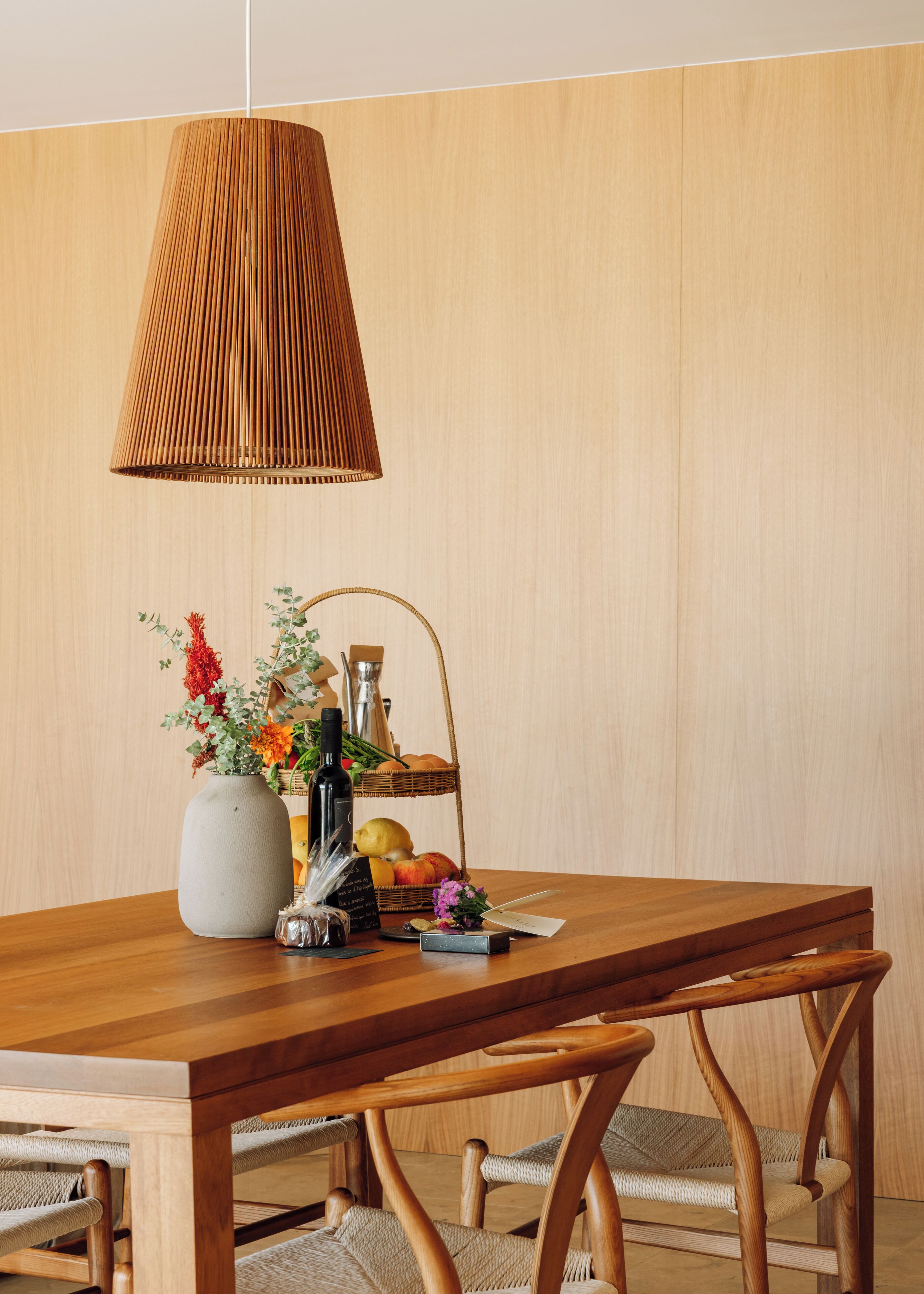 Una mesa de comedor de madera con una lámpara colgante tejida, un florero con flores, una cesta de frutas y una botella.