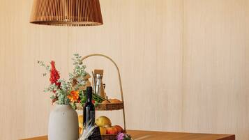 Una mesa de comedor de madera con una lámpara colgante tejida, un florero con flores, una cesta de frutas y una botella.