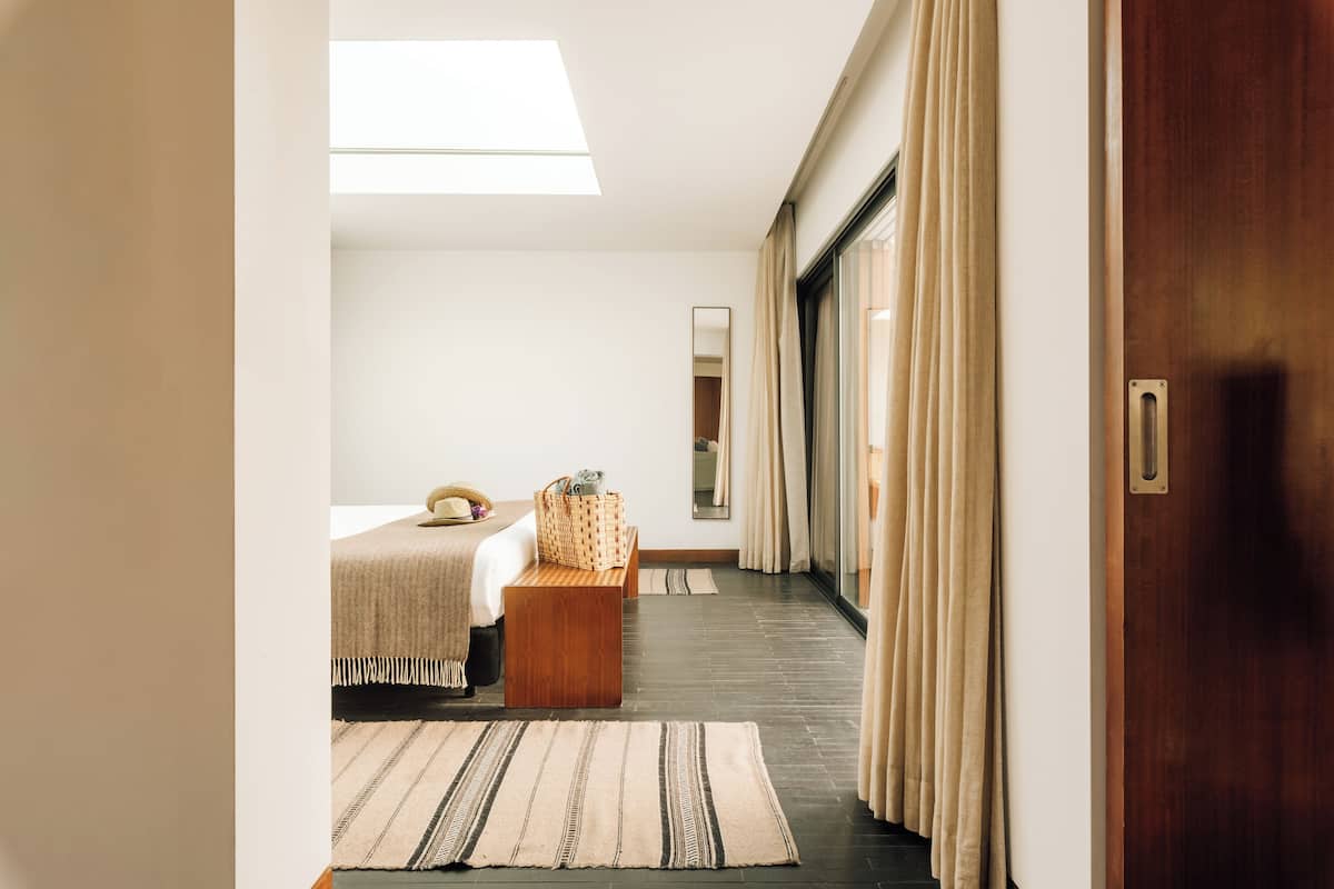 A hotel room with a bed, a wooden bench, a large mirror, and a sliding glass door.