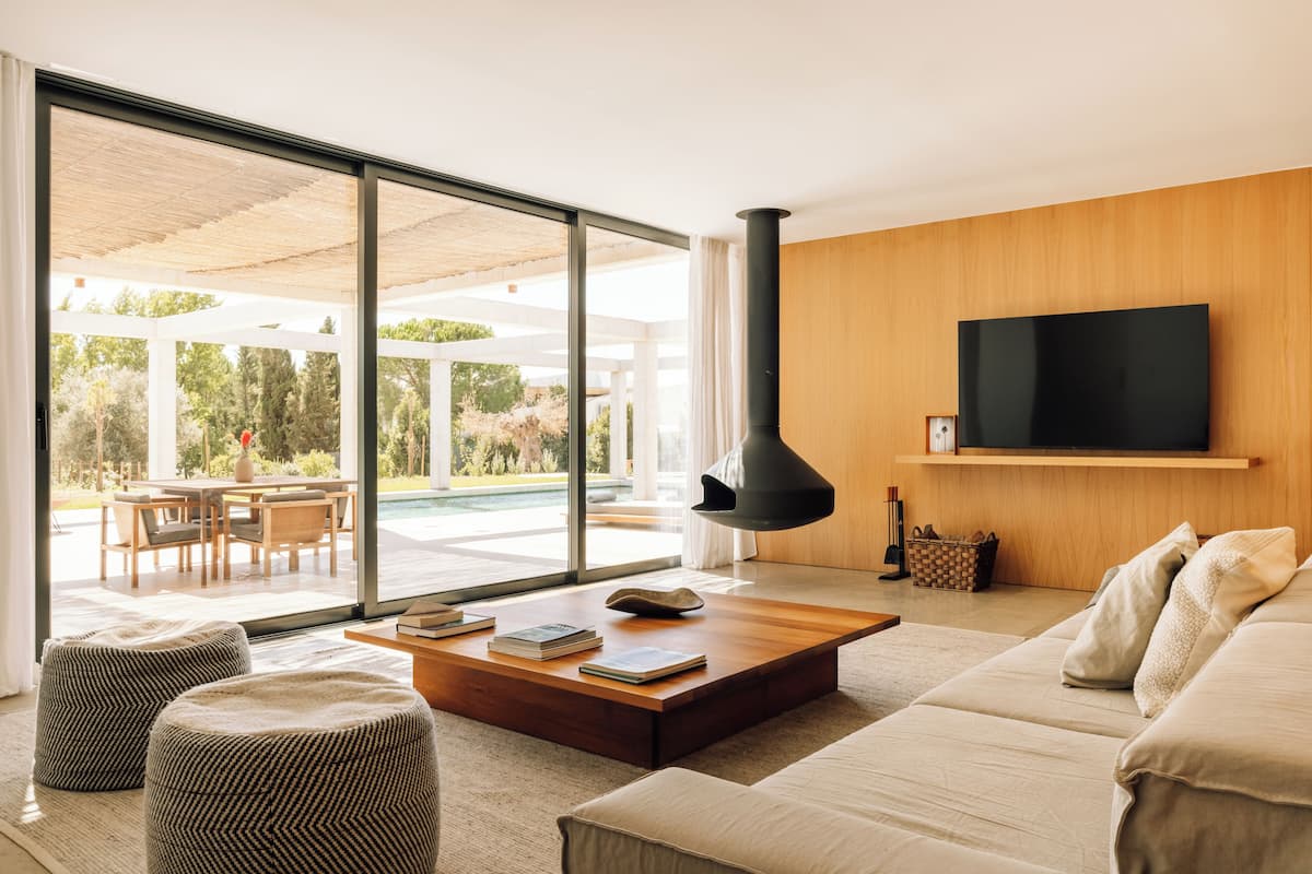 A modern living room with a large sliding glass door leading to an outdoor patio, a wooden coffee table with books, a sofa, and a flat-screen TV mounted on the wall.