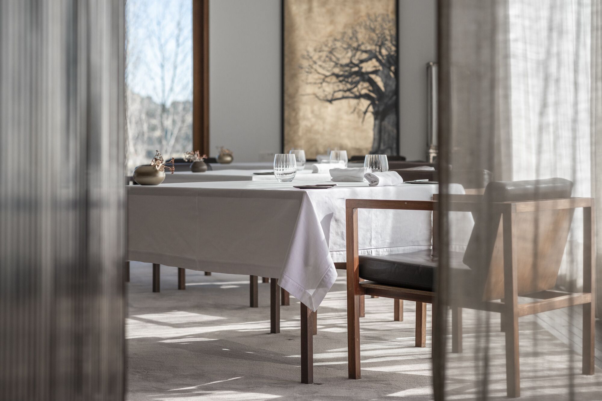 Un comedor con una mesa larga dispuesta para dos personas, con mantel blanco, sillas de madera y un jarrón con flores secas.