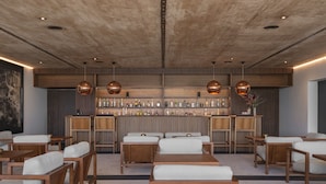 Een bar met houten planken en hangende koperen hanglampen. Er zijn verschillende zithoeken met houten tafels en witte, beklede stoelen.