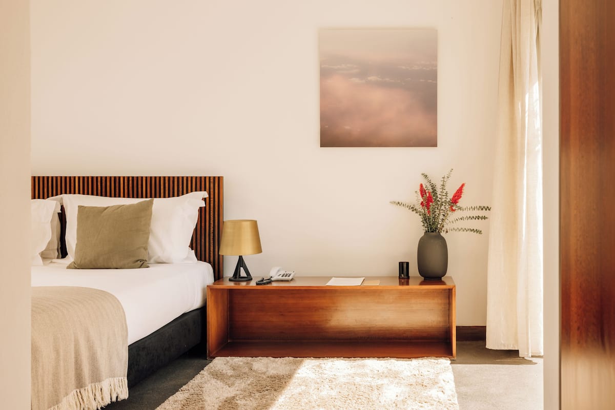A hotel room with a bed, bedside table, lamp, vase with flowers, and a painting on the wall.