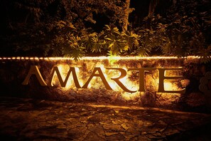 A sign with the word "AMARTE" illuminated by lights, set against a backdrop of tropical foliage and a stone wall.