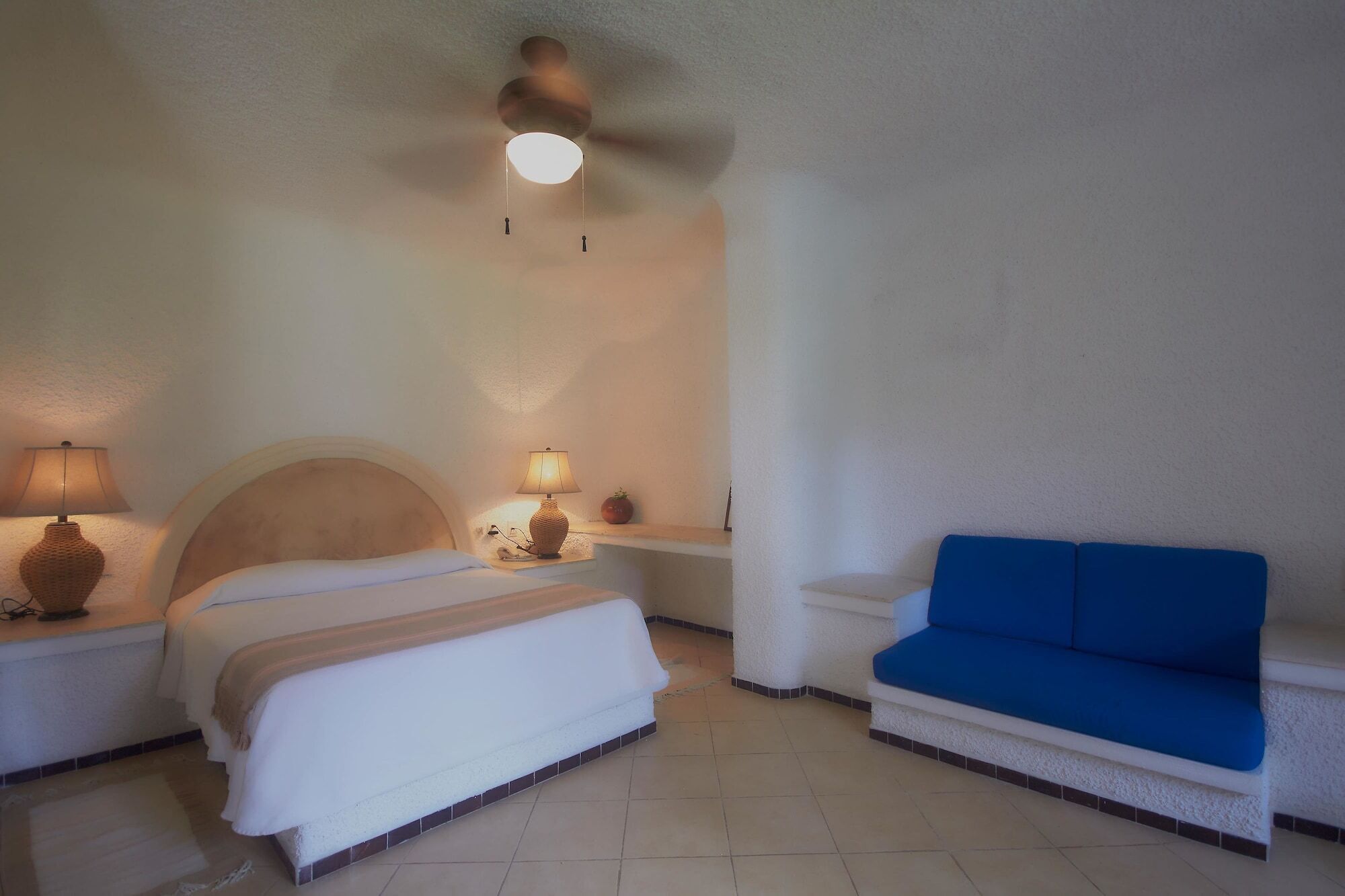 A bedroom with a bed, a nightstand, a lamp, a blue sofa, and a ceiling fan.