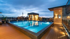 En pool på taket med ett trädäck, en liten byggnad med belysning och utsikt över stadens silhuett.