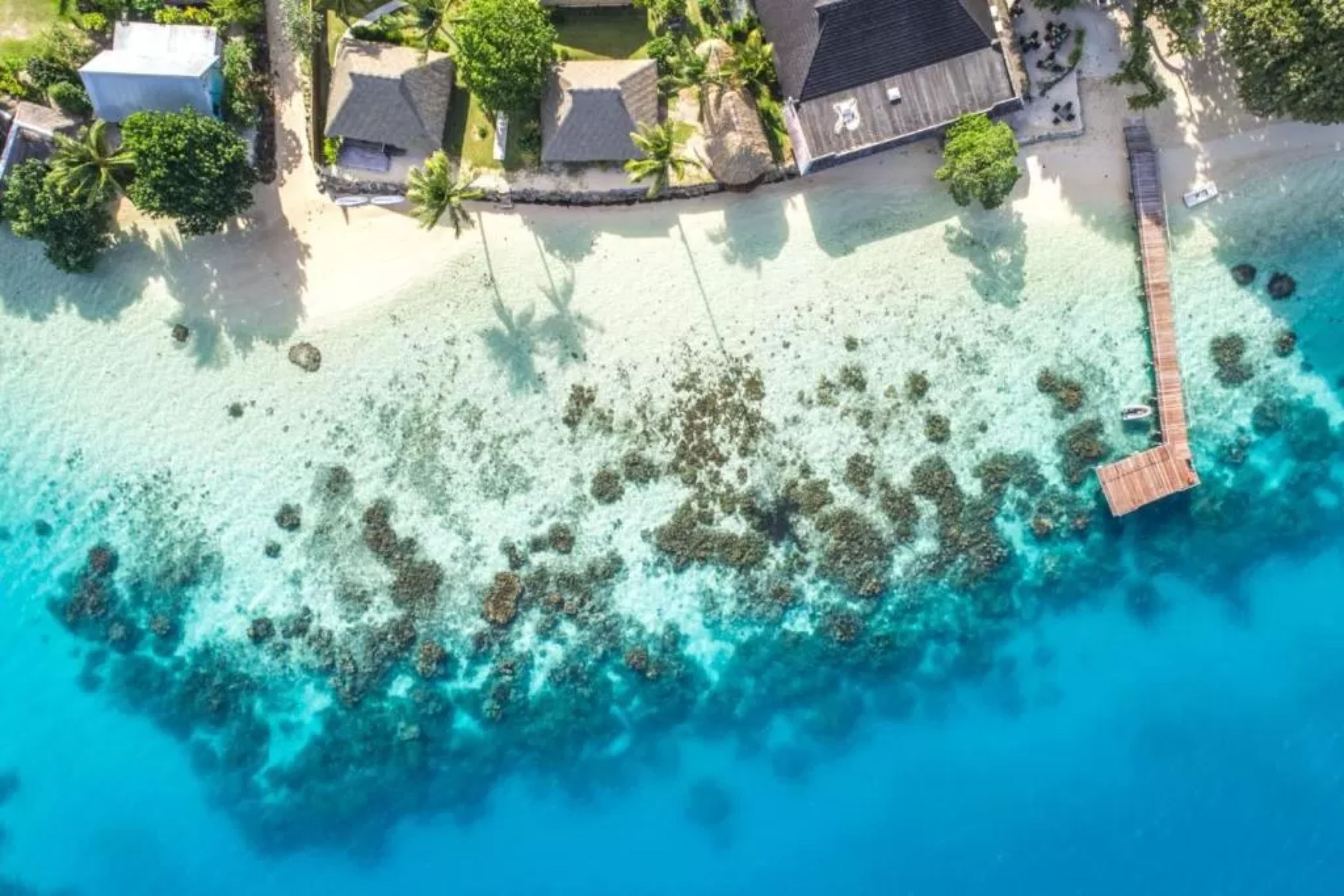 Luchtbeeld van een resort met een strand, helder turquoise water en een houten pier.
