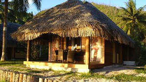 Una cabaña con techo de paja y fachada de madera, ubicada en una playa arenosa con palmeras al fondo.