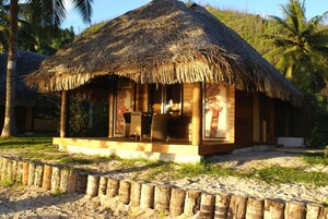Una cabaña con techo de paja y fachada de madera, ubicada en una playa arenosa con palmeras al fondo.