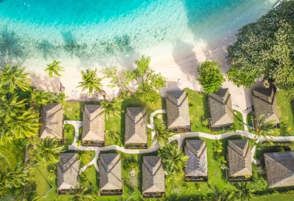Vista aérea de un resort con alojamientos tipo bungalow alineados a lo largo de una playa, conectados por senderos y rodeados de exuberante vegetación y aguas turquesas cristalinas.