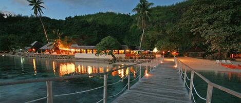 A wooden boardwalk leading to a beachfront resort with palm trees and a mountain backdrop.