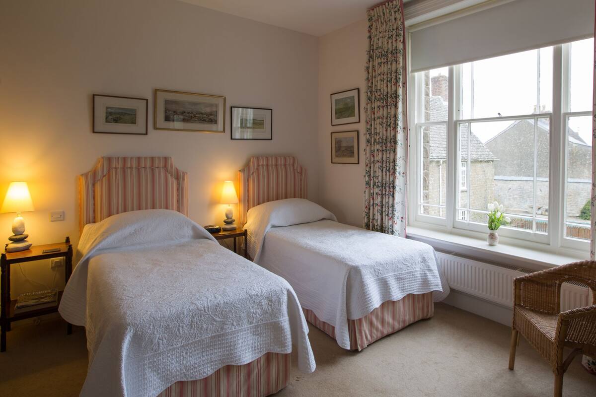 A hotel room with two single beds, a nightstand with a lamp, a chair, a window with curtains, and framed pictures on the wall.