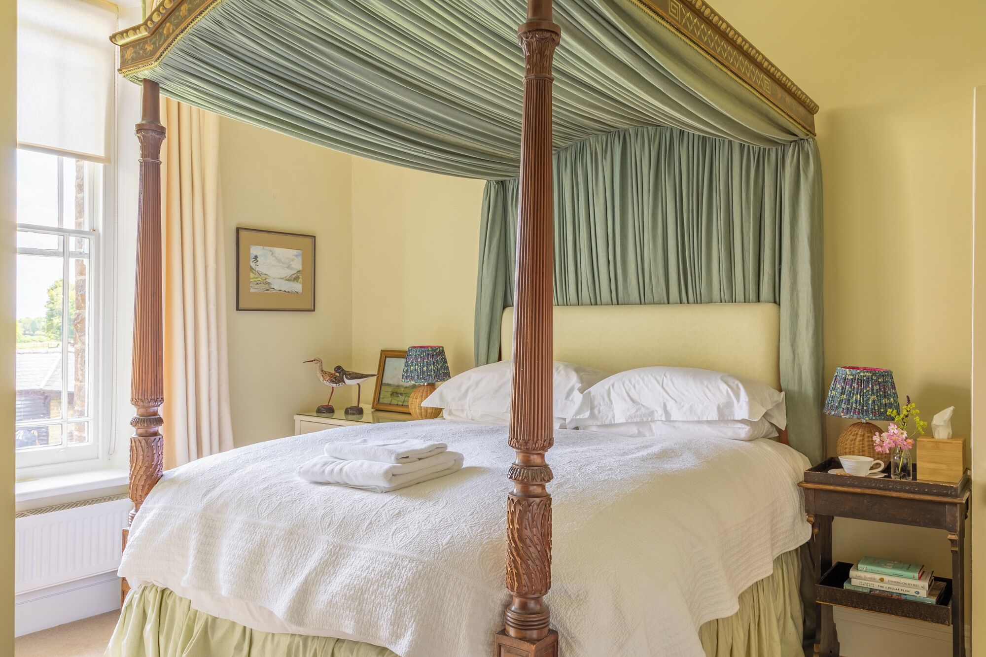 A four-poster bed with a green canopy, white linens, and a bedside table with a lamp and books.