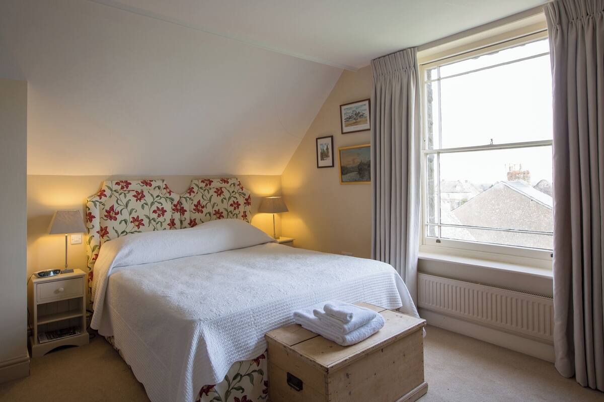 A bedroom with a bed, two bedside lamps, a nightstand, a window with curtains, and a radiator.