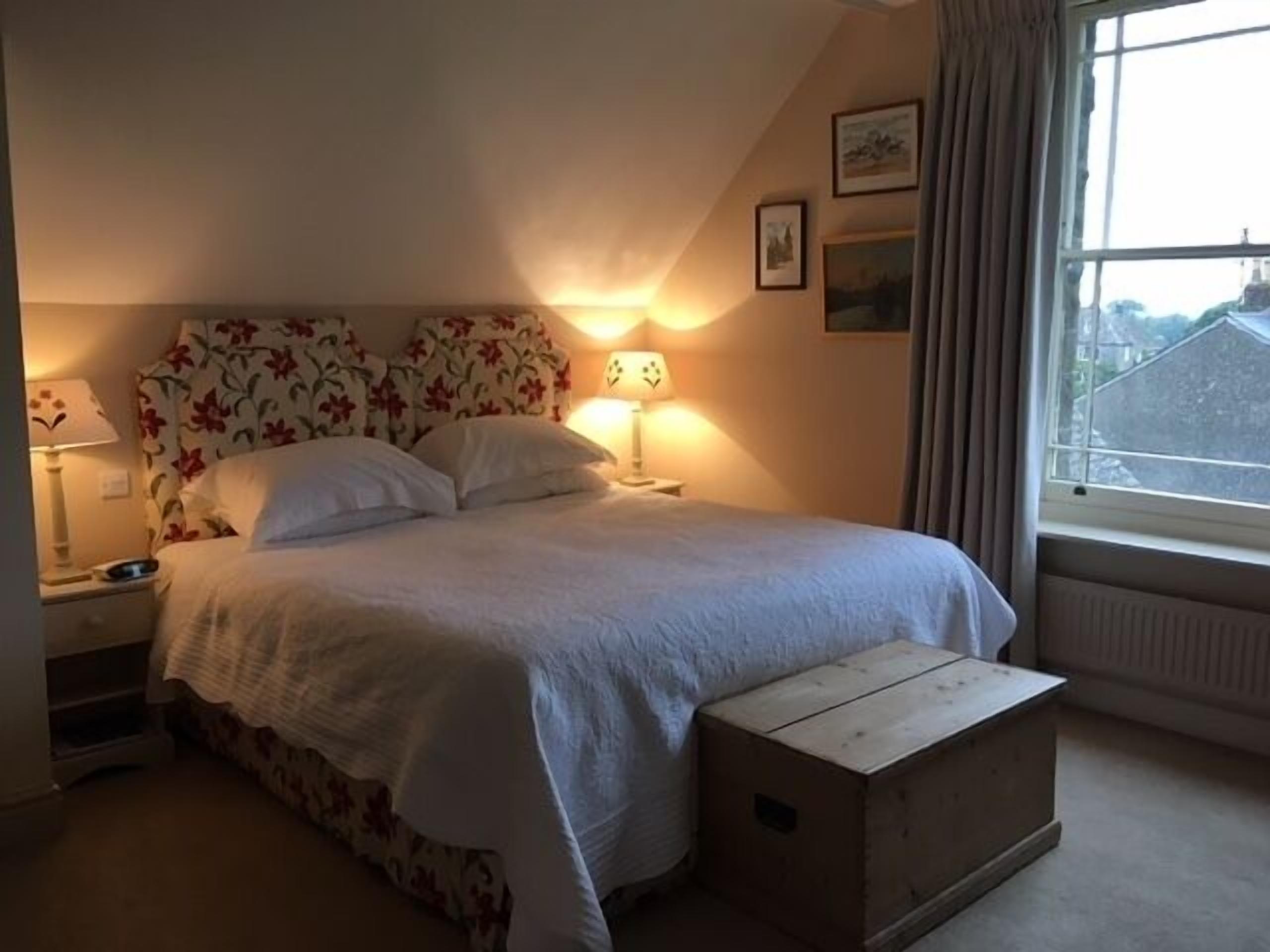 A bedroom with a bed, two bedside lamps, a window with curtains, and a radiator.