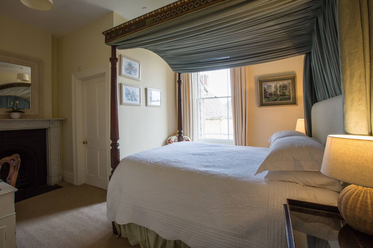 A traditional bedroom with a four-poster bed, a fireplace, and framed artwork on the walls.