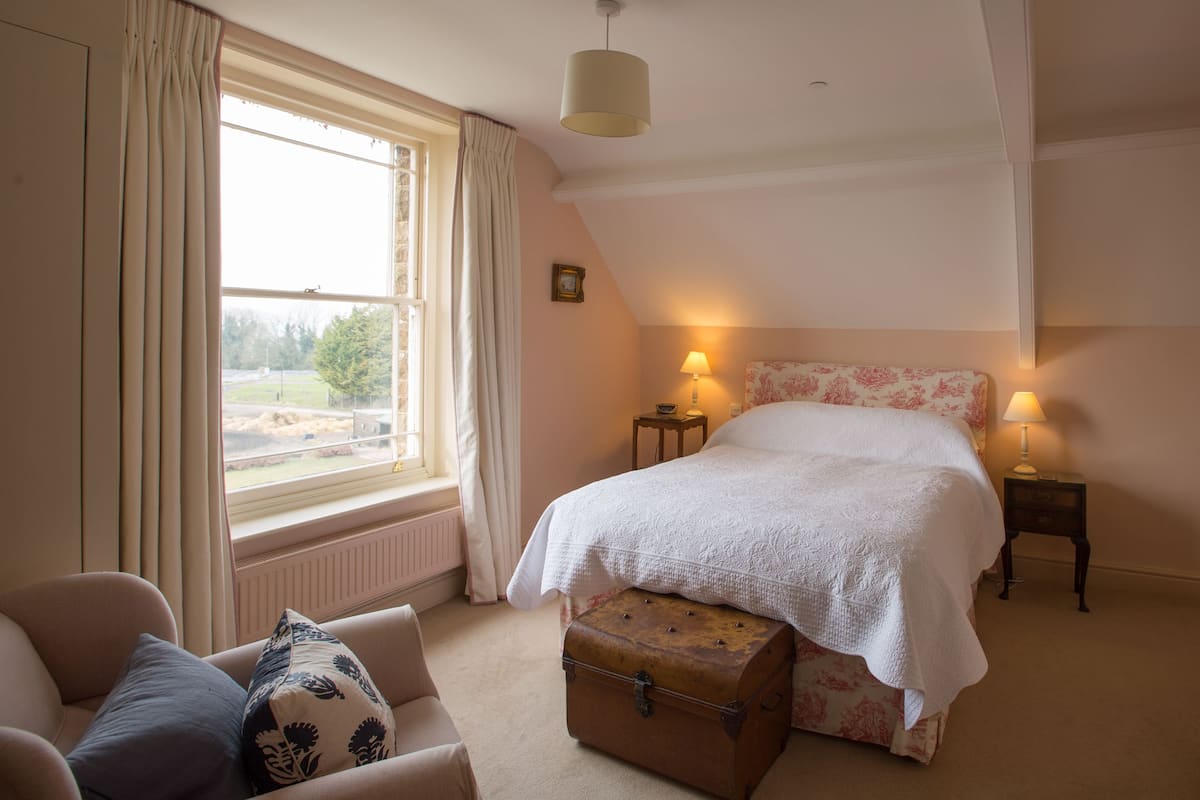 A bedroom with a large bed, two bedside tables with lamps, a window with curtains, a radiator, a chest of drawers, and a chair.