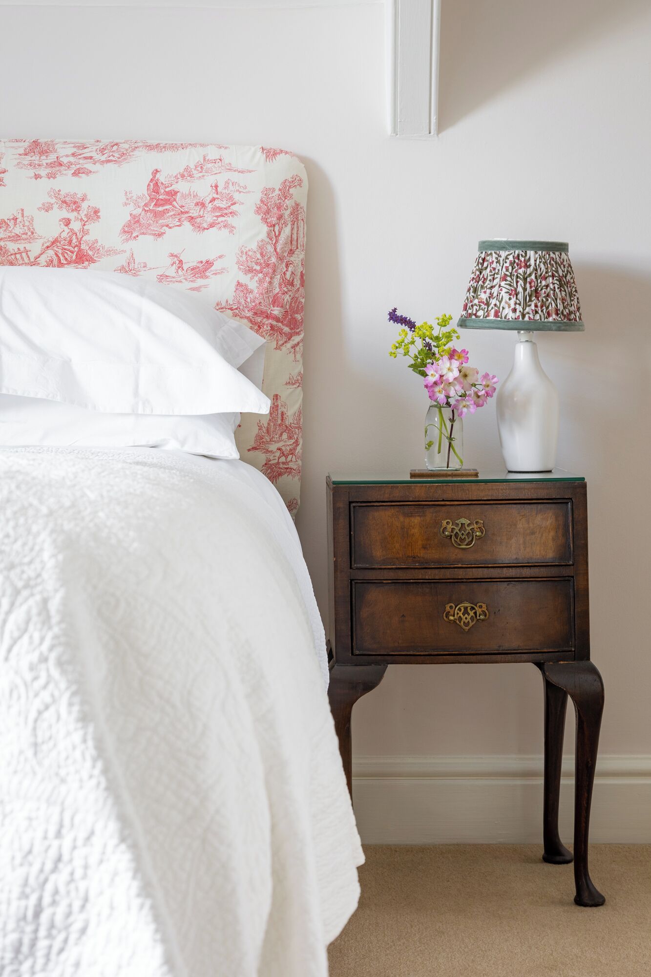 A bedroom with a bed, a nightstand with a lamp, and a vase with flowers.