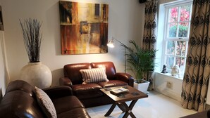 A living room with a brown leather sofa, a coffee table, a large abstract painting, a vase with tall plants, and a window with curtains. - Glangwili Mansion (Carmarthen)