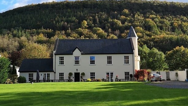 A large, white building with a steep roof and a prominent tower, surrounded by a well-maintained lawn and a forested hill in the background. - Glangwili Mansion (Carmarthen)