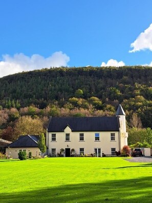 A large, single-story building with a prominent tower, surrounded by a well-maintained lawn and a forested hill in the background. - Glangwili Mansion (Carmarthen)