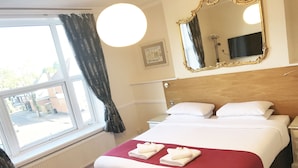A hotel room with a large bed, a bedside table, a nightstand, a chair, a window with curtains, a wall-mounted TV, and a framed picture. - Gidea Park Hotel (Romford)