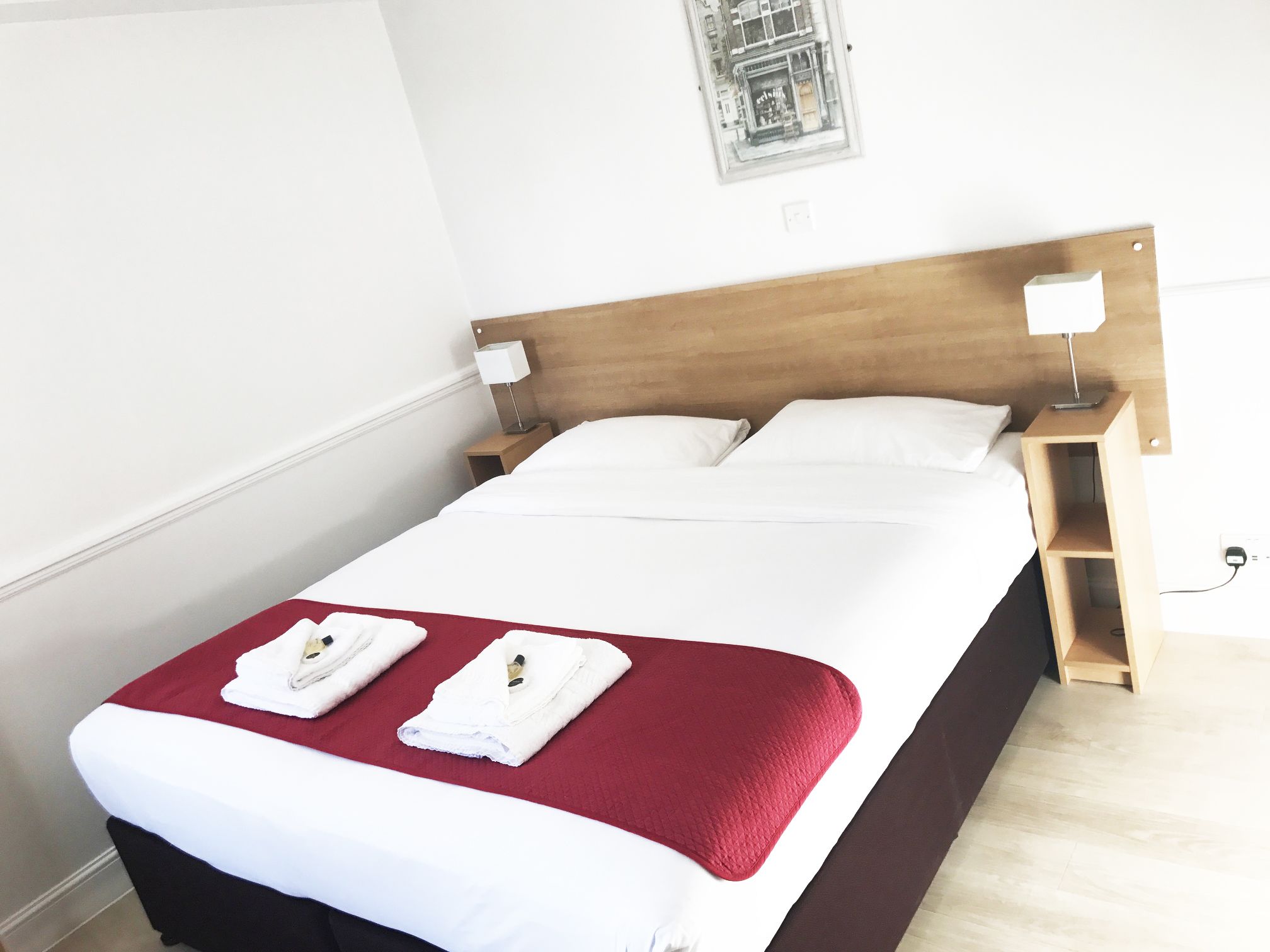 A neatly made bed with white linens and a red blanket, two bedside tables with lamps, and a small window with a view of the street.