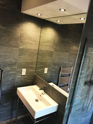 A modern bathroom with a rectangular sink, a glass shower enclosure, and dark tiled walls. - Gidea Park Hotel (Romford)