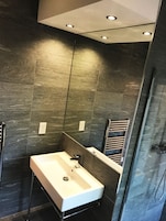 A modern bathroom with a rectangular sink, a glass shower enclosure, and dark tiled walls.