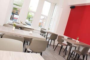 A modern dining area with multiple tables and chairs, large windows, and a red accent wall.
