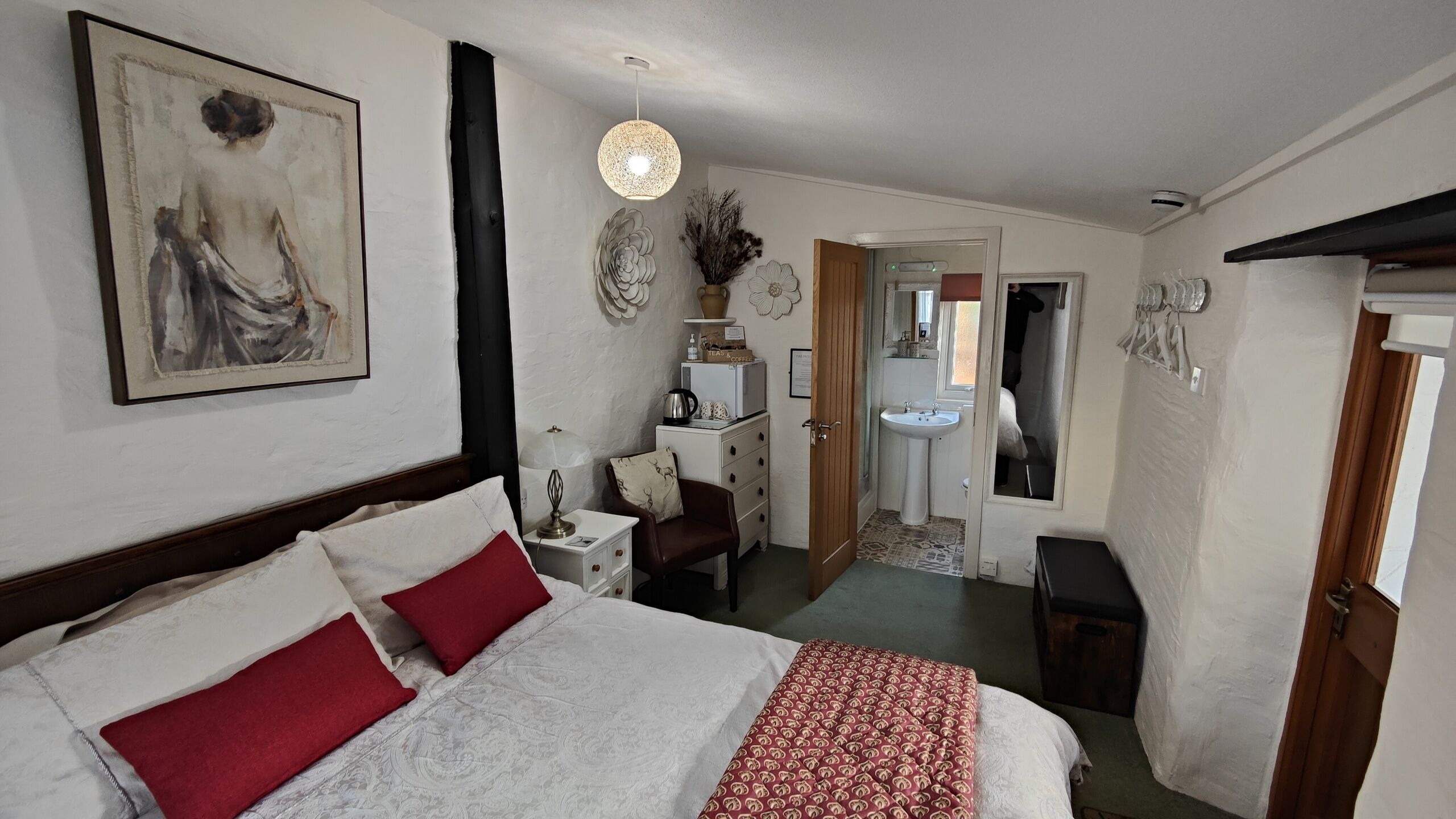 A bedroom with a large bed, a desk, a chair, a dresser, a mirror, and a bathroom visible through an open door.