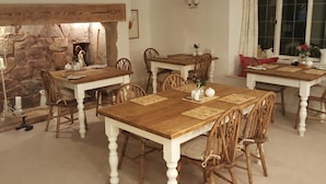 A dining area with wooden tables and chairs, a stone wall, and a window with curtains. - Steps Farmhouse (Minehead)