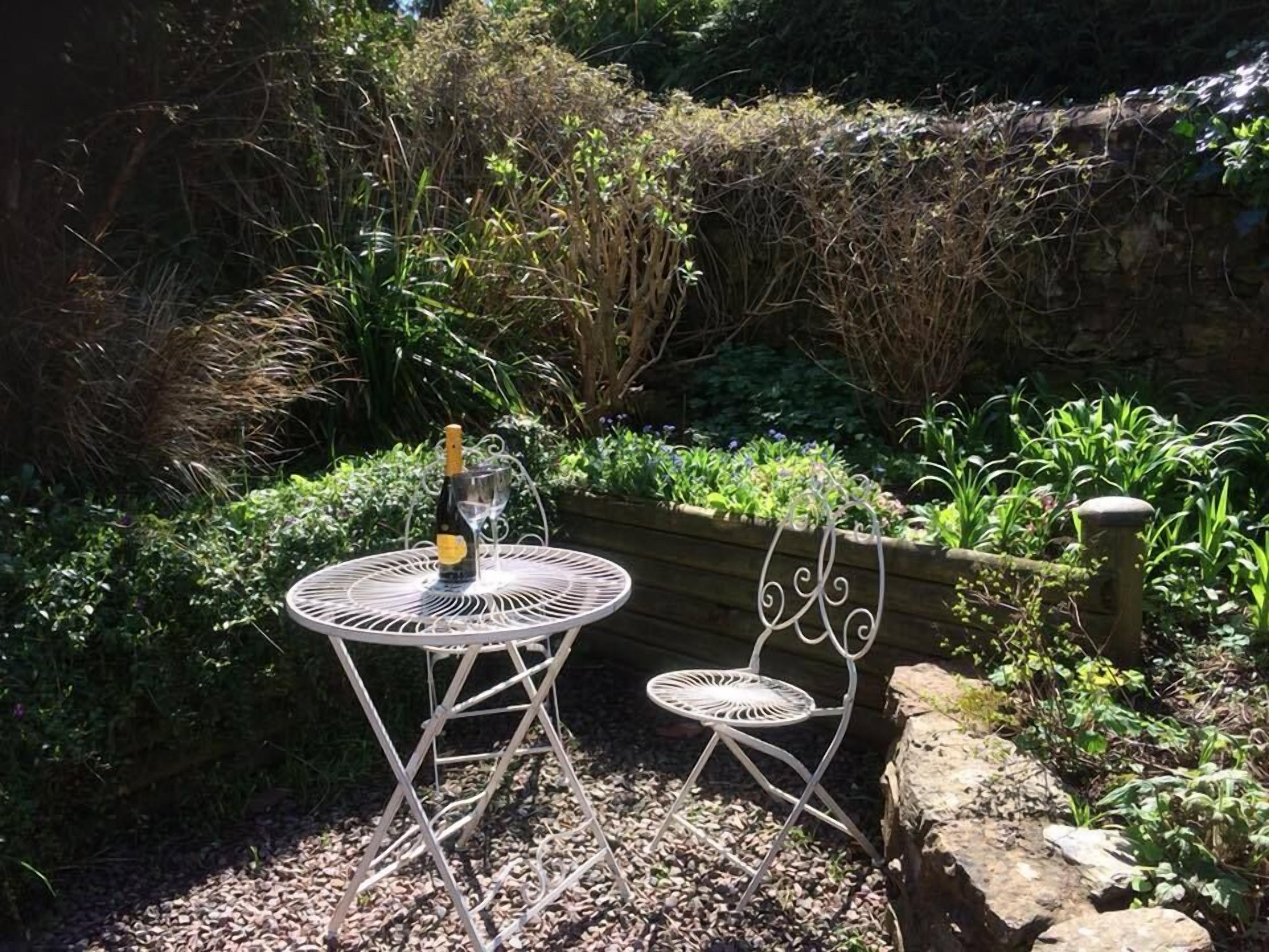 A garden with a metal table and two chairs, a bottle of champagne, and a glass.