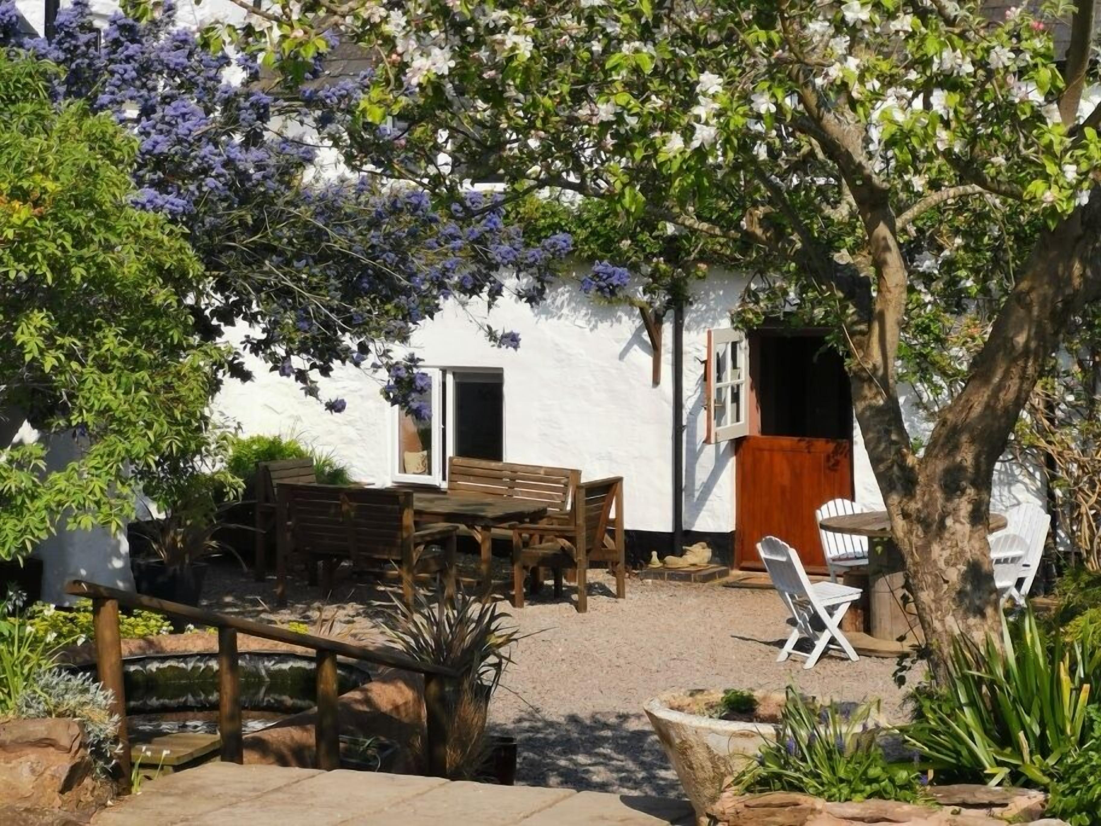 Eine Terrasse mit Holztisch und -stühlen, ein weißes Gebäude mit einer Tür und ein Baum mit violetten Blüten.