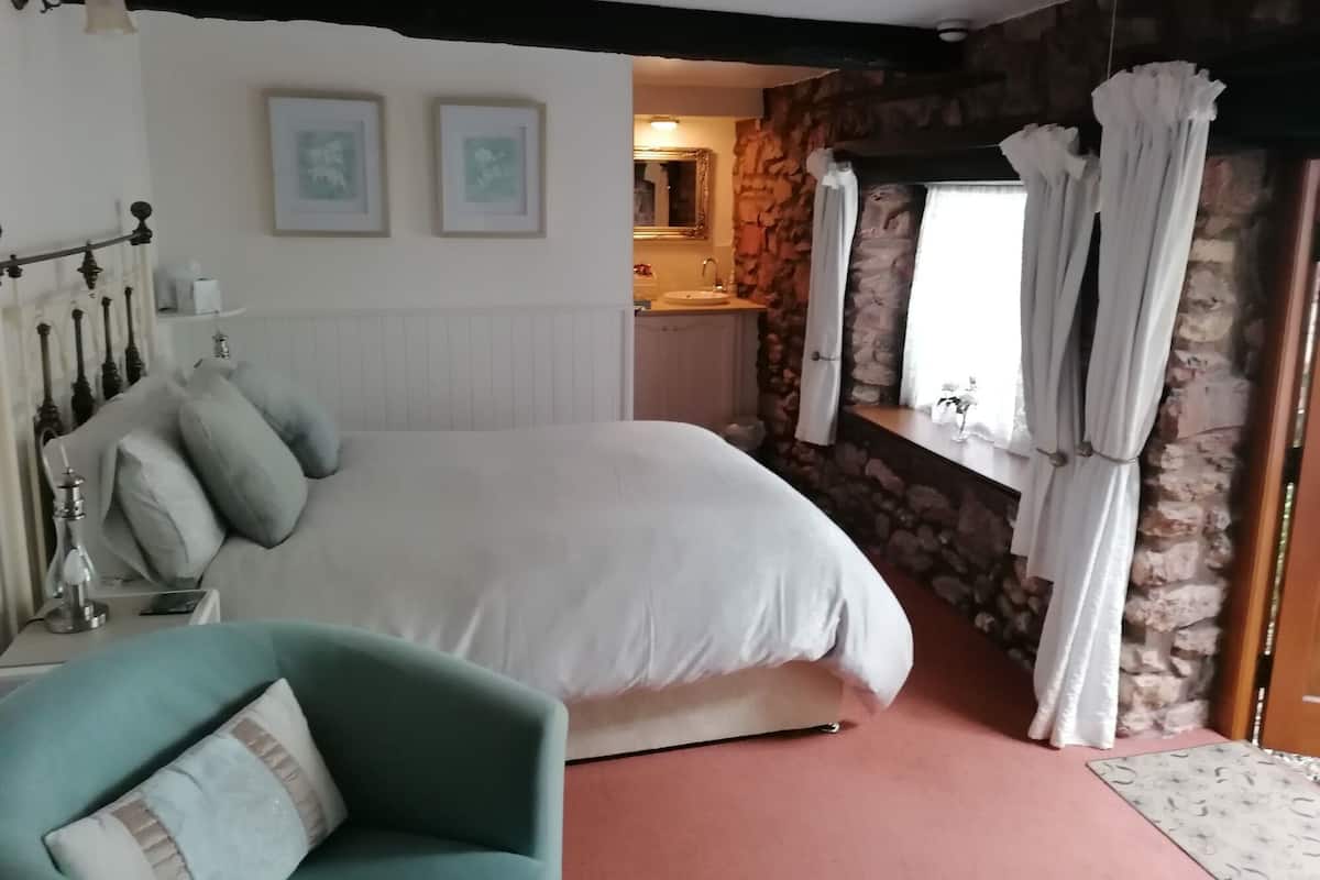 A bedroom with a large bed, a bedside table, a chair, a stone wall, a window with curtains, and a bathroom visible in the background.