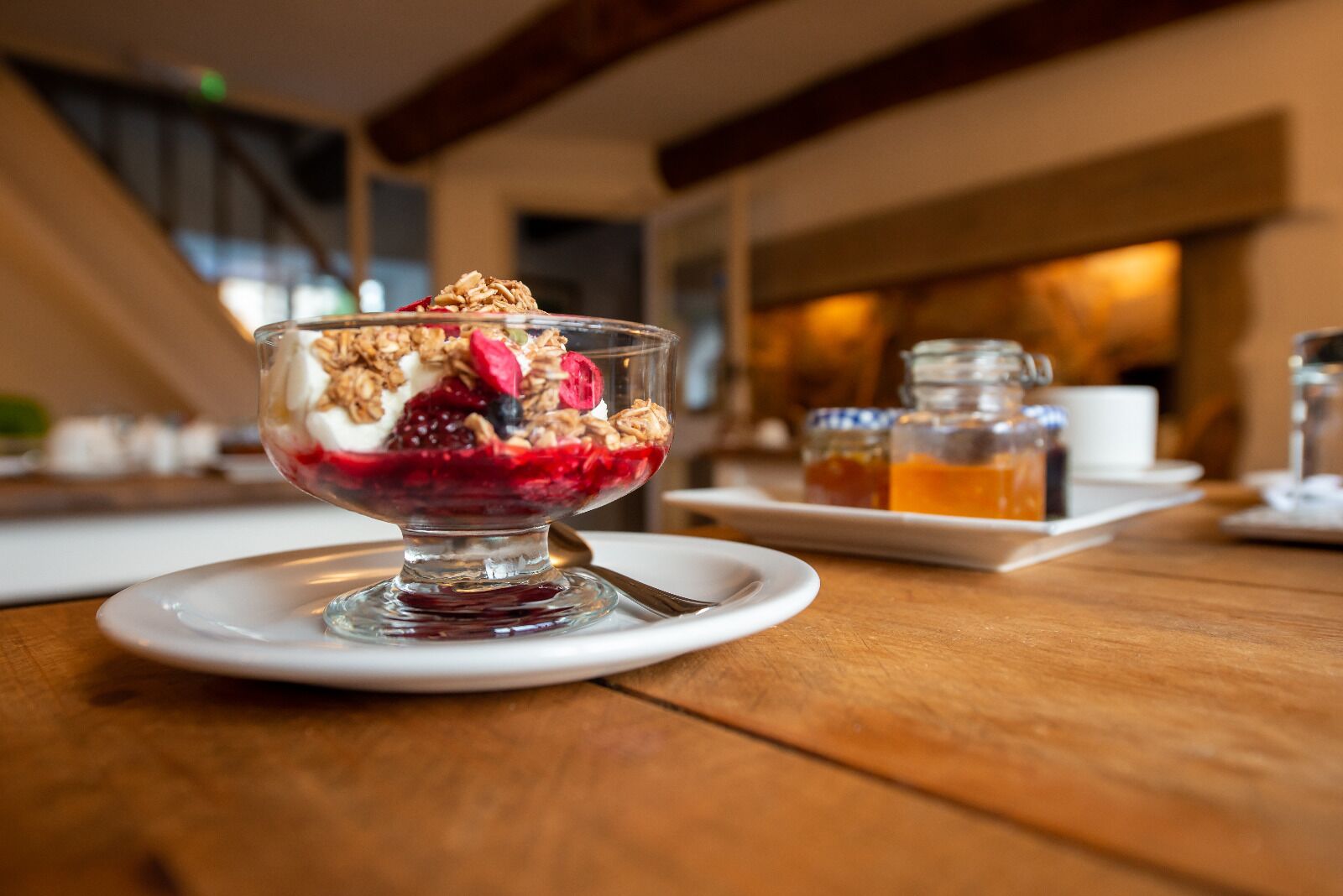 Un postre en un cuenco de vidrio sobre un plato blanco, con una mesa de madera y una taza blanca al fondo.