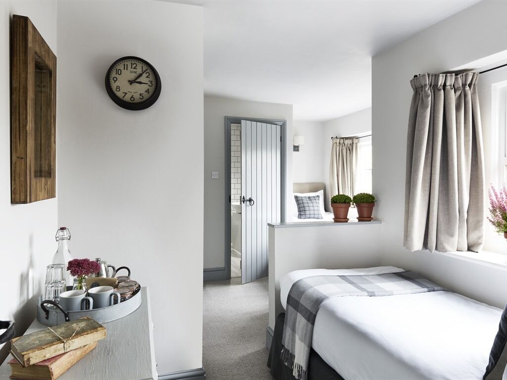 Un dormitorio con cama, mesita de noche con un reloj, ventana con cortinas y una puerta que da a otra habitación.