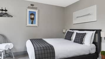 Un dormitorio ordenado con cama, una silla, una mesita con una planta y dos cuadros en la pared.