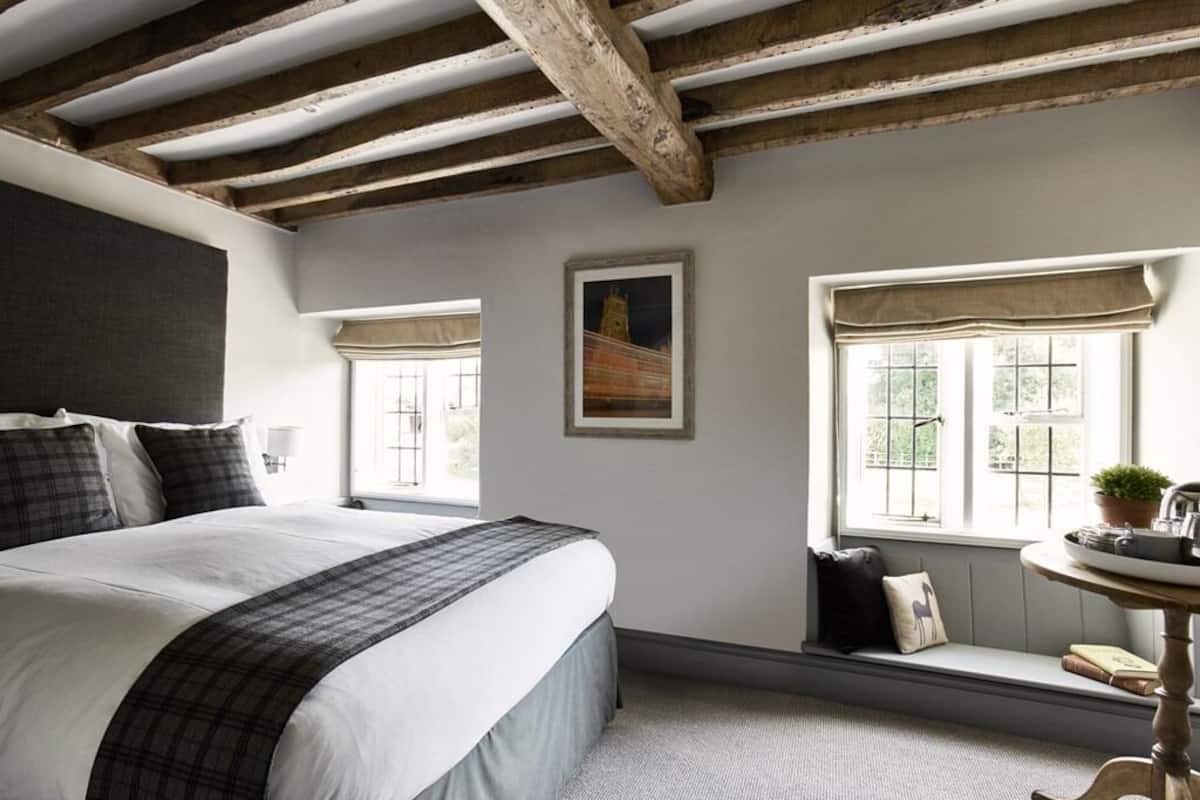 Un dormitorio con una cama grande, dos ventanas, un banco y una mesita con una planta.