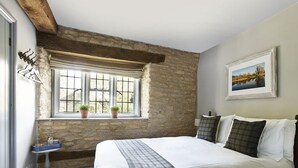 A bedroom with a large bed, a stone wall, a window with plants, and a painting on the wall. - The Masons Arms (Cirencester)