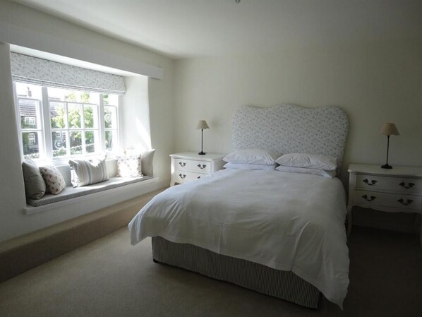 A bedroom with a large bed, two bedside tables with lamps, a window with a view of greenery, and a built-in bench with pillows.