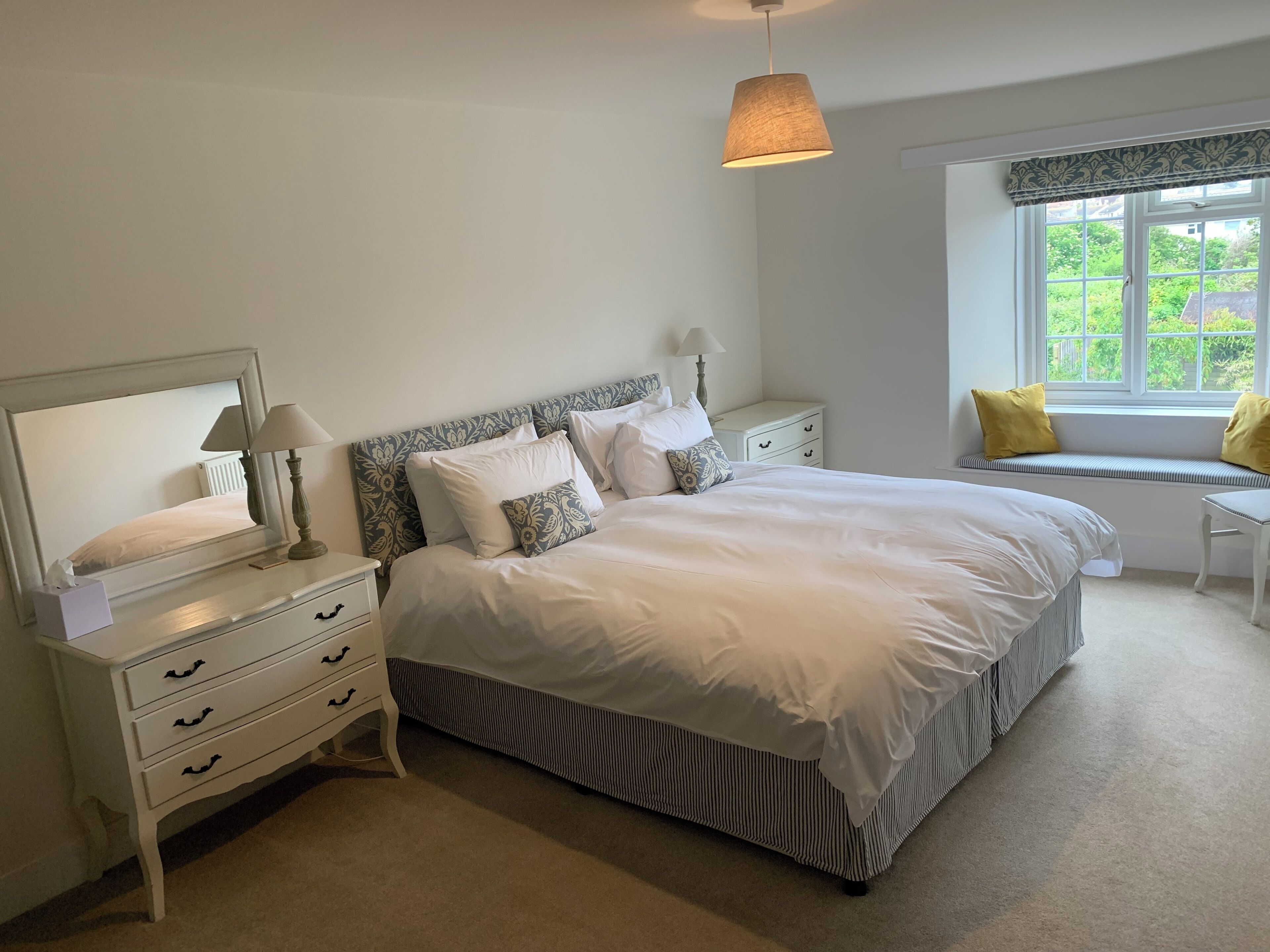 Un dormitorio ordenado con cama, mesitas de noche, ventana con vistas y una lámpara colgante.