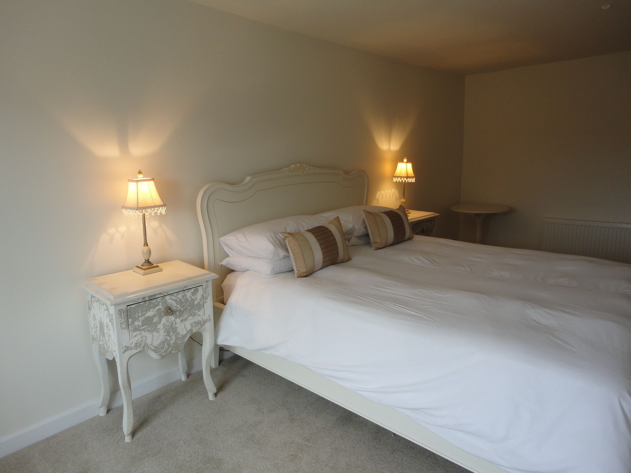A neatly made bed with white linens and striped pillows, flanked by two bedside tables with lamps, one of which has a decorative top.
