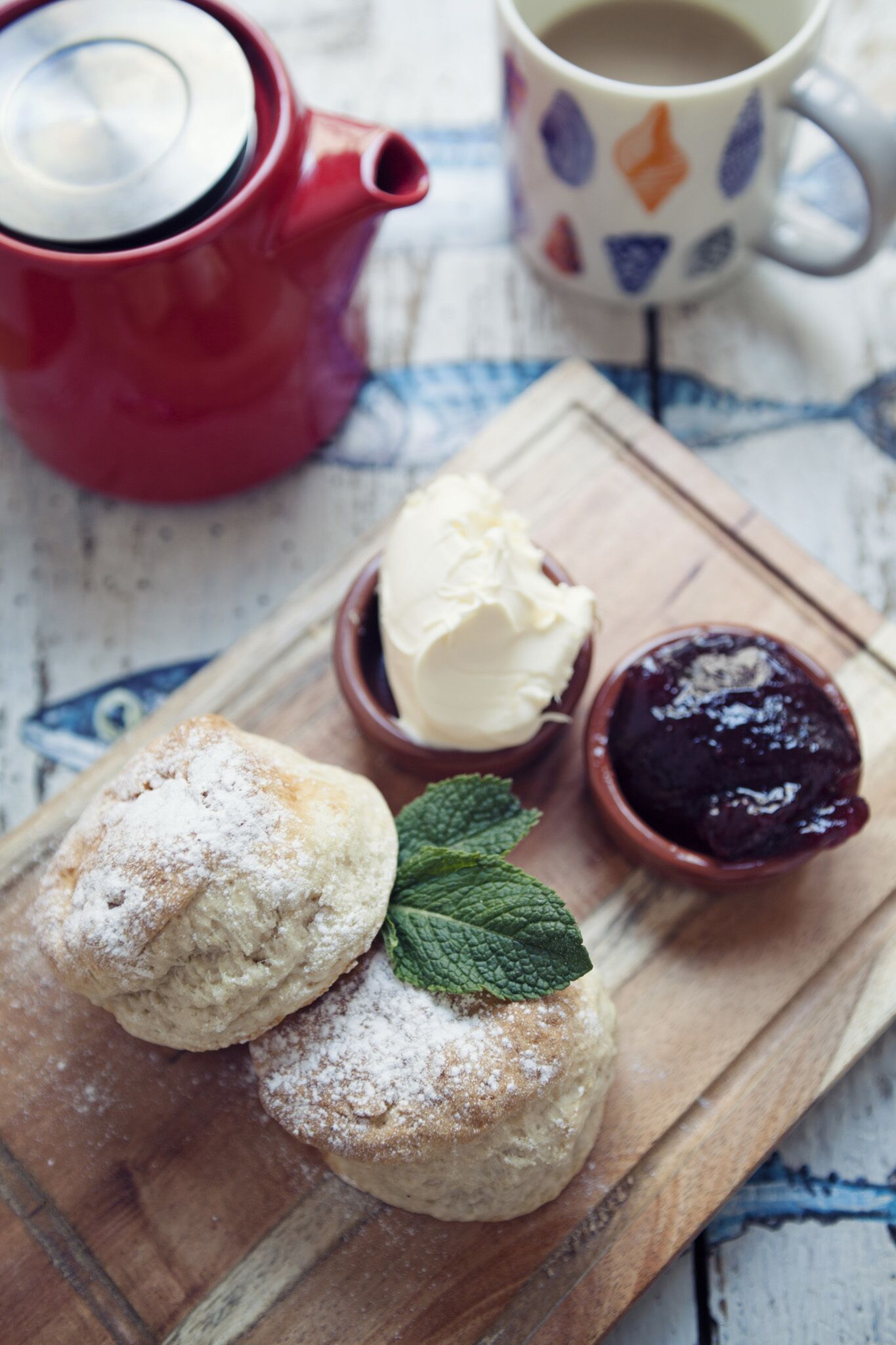 A wooden board with two scones dusted with powdered sugar, a small bowl of butter, and a bowl of jam, accompanied by a red teapot and a patterned mug.