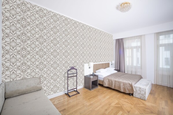 Une chambre d’hôtel avec un papier peint à motifs, un lit, une table de chevet, une commode, une lampe suspendue, un banc et une fenêtre avec des rideaux.