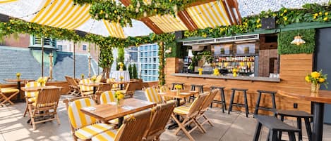 An outdoor seating area with tables and chairs, a bar, and greenery.