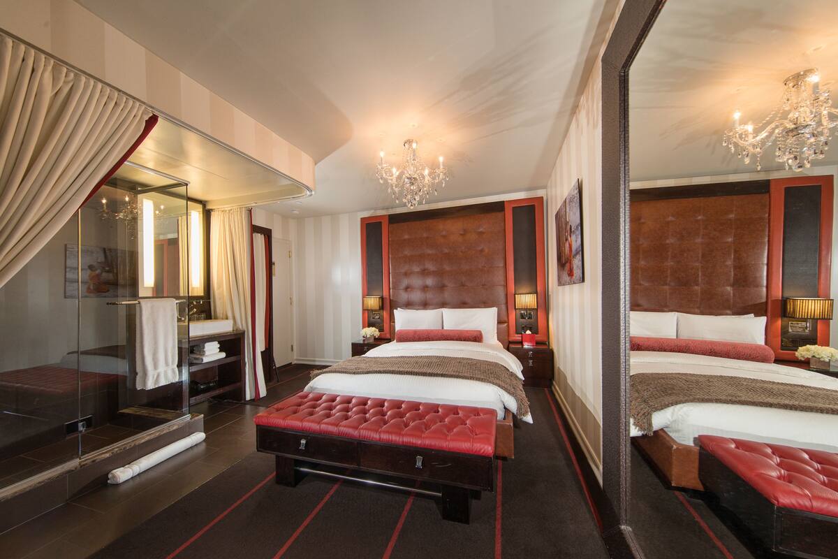 A hotel room with two beds, a large mirror, a red upholstered bench, and a chandelier.