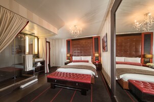 Una habitación de hotel con dos camas, un espejo grande, un banco tapizado en rojo y un candelabro.