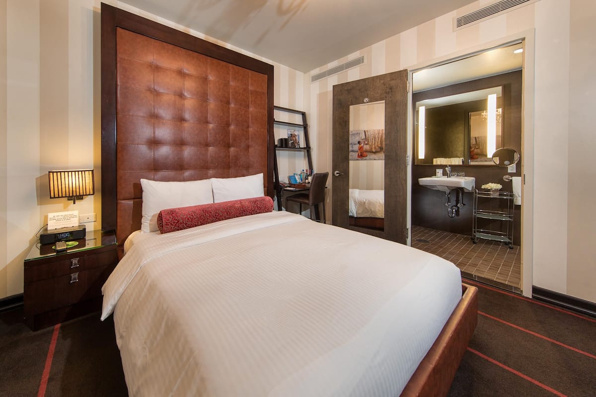 A hotel room with a large bed, a bedside table with a lamp, a wardrobe, and a bathroom visible through an open door.