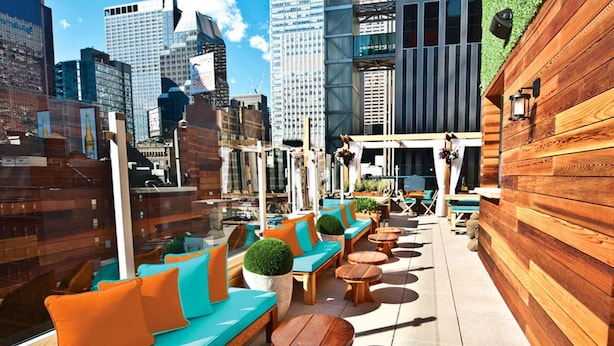 A rooftop terrace with wooden seating and tables, offering a view of the city skyline.